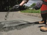 Sable à joints par Colas : Réparation ponctuelle de chaussée : agent en EPI répandant un enrobé à froid pour reboucher un nid-de-poule en bordure de route.