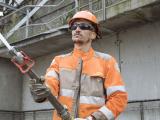 Collaborateur de Premys, filiale de Colas : Ouvrier de chantier en tenue de sécurité (casque et vêtement haute visibilité) manipulant un flexible / un tuyau d’intervention dans un environnement industriel en ossature métallique, illustrant sécurité opérationnelle, intervention sur réseau et maîtrise des gestes professionnels.