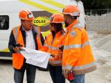 Trois collaborateurs en tenue de chantier consultant des plans devant un véhicule COLAS, illustrant coordination, expertise technique et pilotage opérationnel.