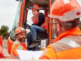 Discussion de chantier entre opérateurs en tenue haute visibilité près d’un engin de terrassement, avec plans et consignes partagés.