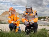 Portrait de collaborateurs Colas : Binôme terrain en EPI (casques, gilets haute visibilité, gants) échangeant sur un chantier d’infrastructure près d’un talus et d’une voirie, illustrant coordination, sécurité et suivi des travaux.