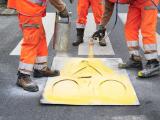 Réalisation piste cyclable provisoire CD94 - AXIMUM : Marquage au pochoir d’un pictogramme vélo en peinture jaune sur chaussée, réalisé par une équipe en EPI orange.