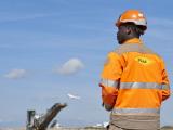 Agent de chantier équipé d’EPI (casque, gilet haute visibilité, radio), sur site aéroportuaire avec engin de chantier au loin, illustrant sécurité, coordination opérationnelle et gestion d’infrastructure.