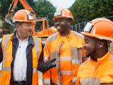 Briefing de sécurité sur chantier routier avec équipe en EPI orange et engins de terrassement en arrière-plan.