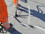 Agent en tenue haute visibilité appliquant un liant et saupoudrant un matériau granulaire pour réparer une fissure d’enrobé sur la chaussée — cadrage rapproché sur les jambes, l’équipement et le produit en action.