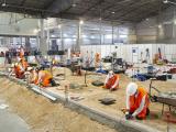  Équipe d'ouvriers en gilets orange et casques travaillant à l'intérieur d'un hall sur des aménagements routiers et voiries en cours d'implantation, démontrant coordination, sécurité et savoir‑faire technique.