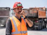 Plateforme Valormat de Trouillas : Chef de chantier en EPI (casque rouge, gilet haute visibilité) coordonnant les opérations par radio devant des camions-bennes, illustrant sécurité, communication et pilotage logistique sur site.