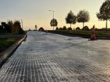Pose de géogrille sur chaussée avant enrobés : vue ras du sol au crépuscule, trame blanche tendue sur la voie, cônes de chantier et alignement d’arbres le long de la route.