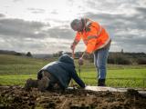 Accompagnement plantation en milieu agricole : encadrant en gilet orange guide un jeune qui positionne un plant à travers un film de paillage, terrain boueux et paysage rural en arrière‑plan.
