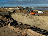Chantier de reprofilage côtier/dragage à l’interface plage‑anse : pelleteuses Liebherr et camions bennes déplacent des volumes de sable, avec clôtures dune/ganivelles et zone portuaire en arrière‑plan.