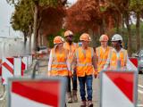 Cinq ouvriers en gilets et casques de haute visibilité traversent un chantier routier balisé le long d’une chaussée bordée d’arbres.
