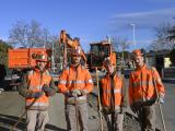 Quatre jeunes collaborateurs en équipement de chantier orange posent avec leurs outils devant des engins de travaux publics sur une route en cours d'aménagement.