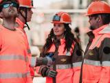 Jeune femme en tenue haute visibilité orange et casque de chantier, souriante sur un chantier de voirie avec des équipes et engins en arrière-plan.